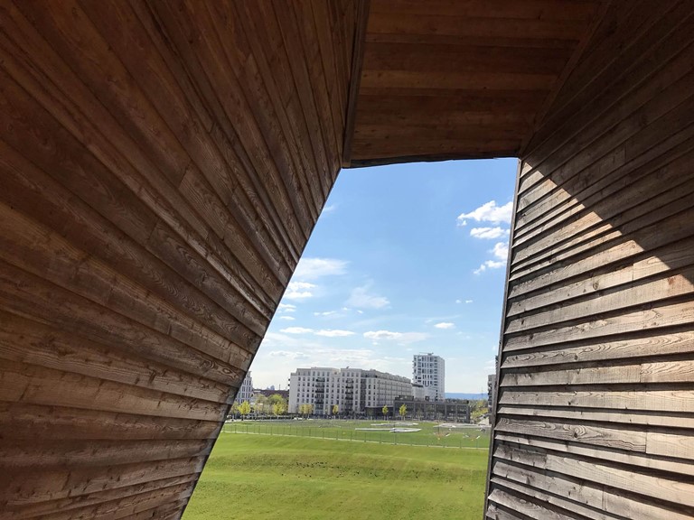 A structure of wooden planks through which one looks at white housing complexes and a light green lawn
