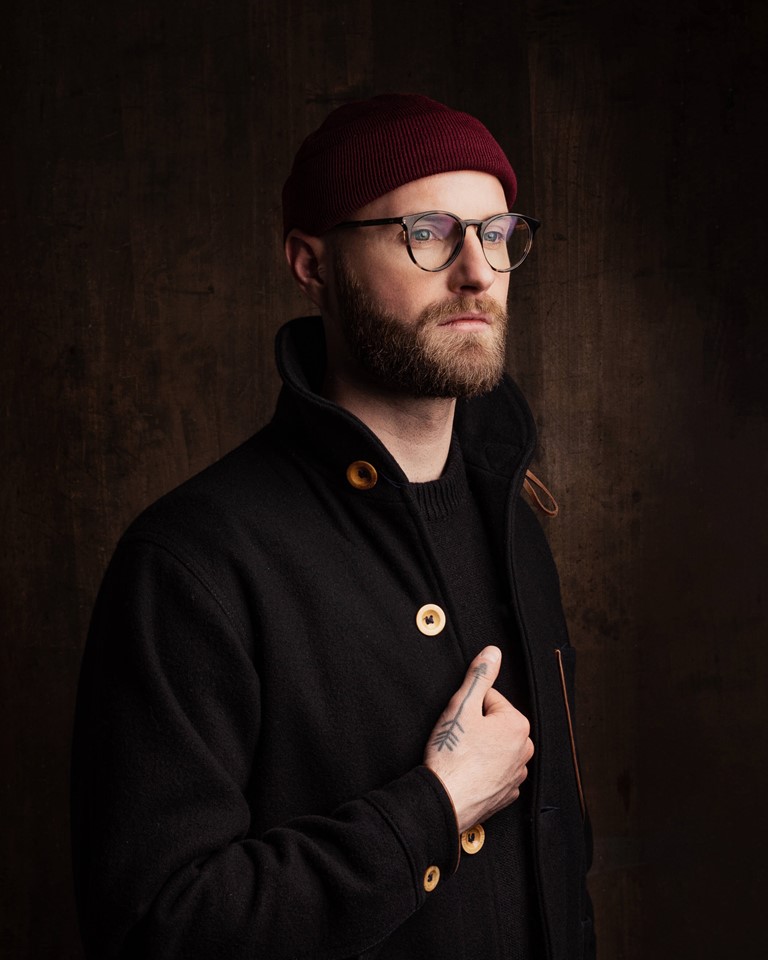 Man with a full beard and horn-rimmed glasses. He is wearing a burgundy knitted cap and a black jacket with large wooden buttons. The right hand on the lapel is decorated with a tattoo in the shape of an arrow.