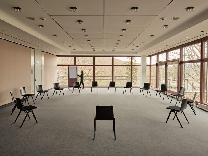 A conference room with floor-to-ceiling windows. Chairs are arranged in a circle in the centre of the room. In the background, a woman is pushing a flipchart.