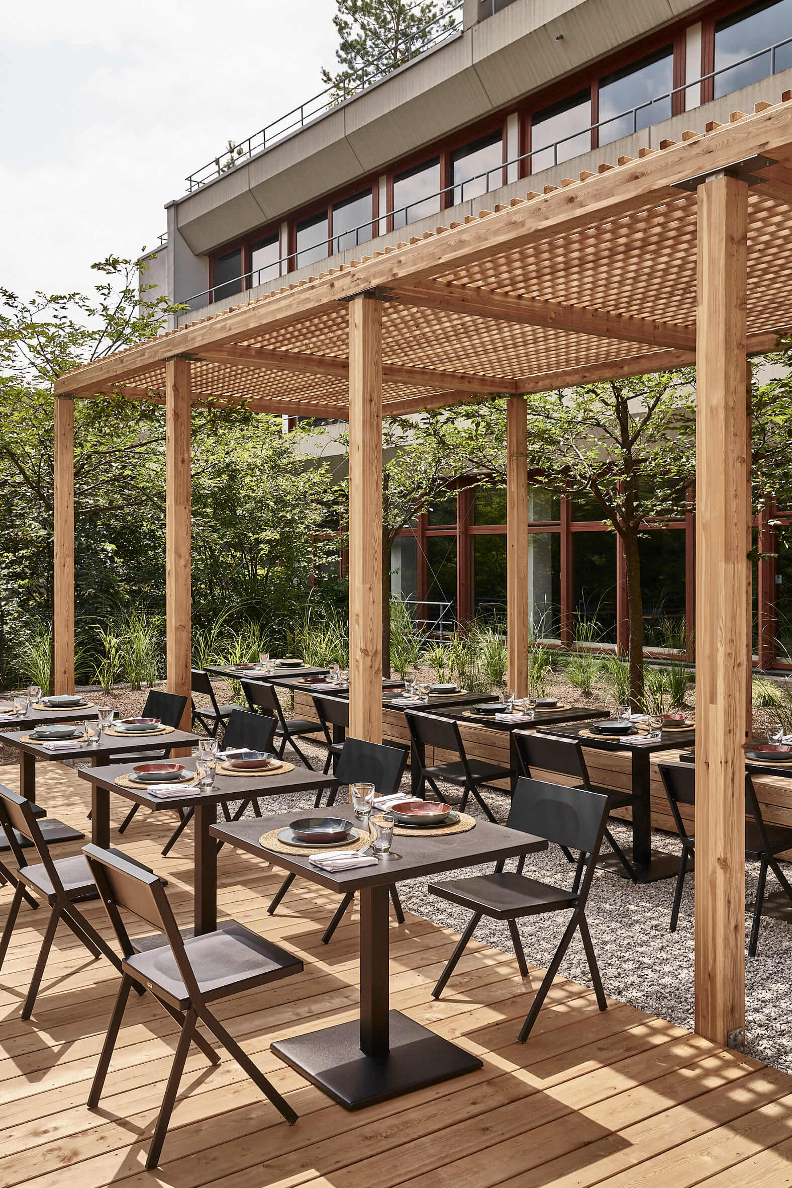 Die atrium terrace im Bold Campus Eingedeckte schwarze Tische und Stühle, die teilweise auf hellen Holzplanken und teilweise im Halbschatten einer hölzernen Pergola stehen.