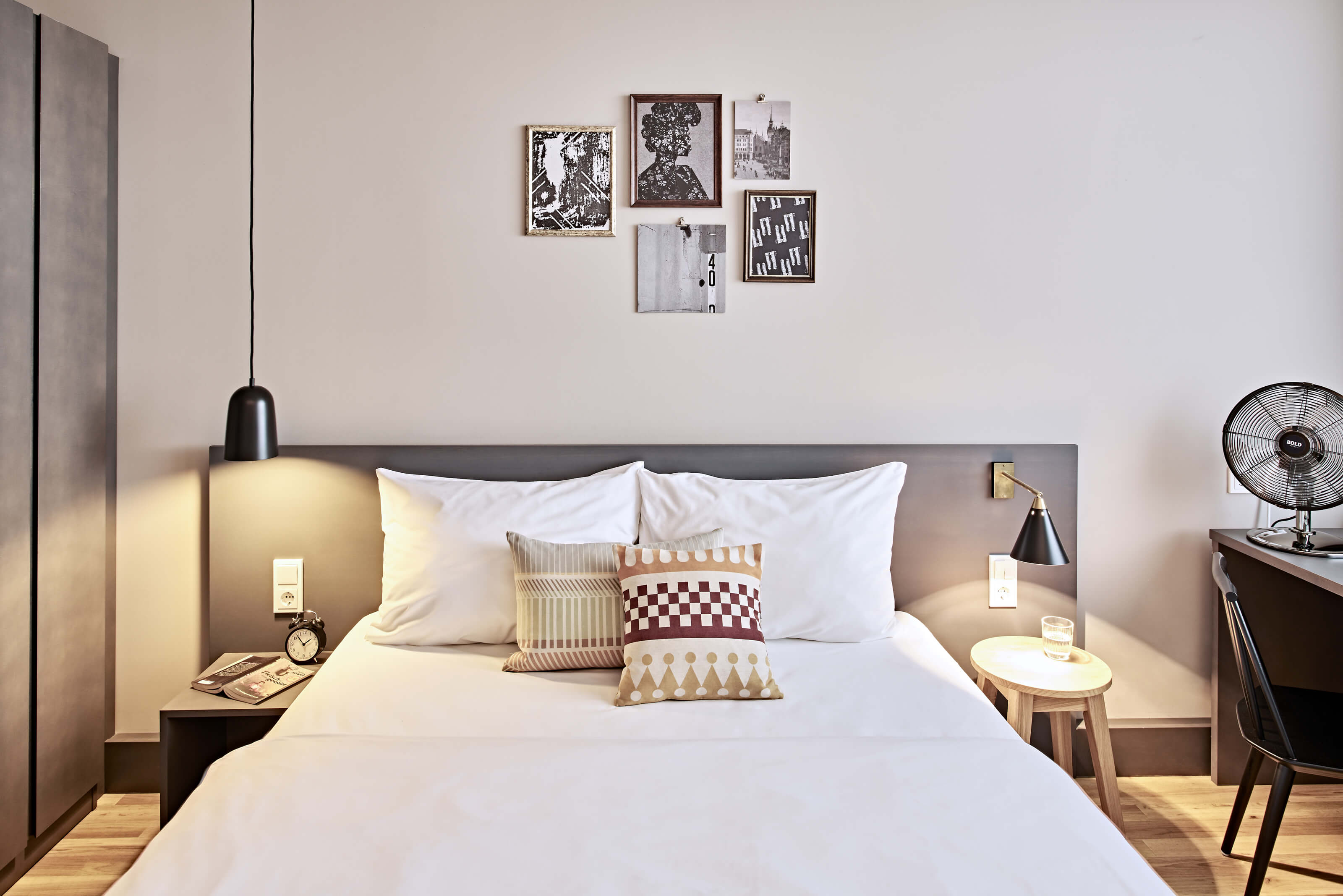 Großes Doppelbett im Bold Room im Bold Hotel München Zentrum Weiß bezogenes Doppelbett auf einem Holzfußboden, links ein grauer Schrank, rechts ein Schreibtisch am Fenster mit Stuhl.