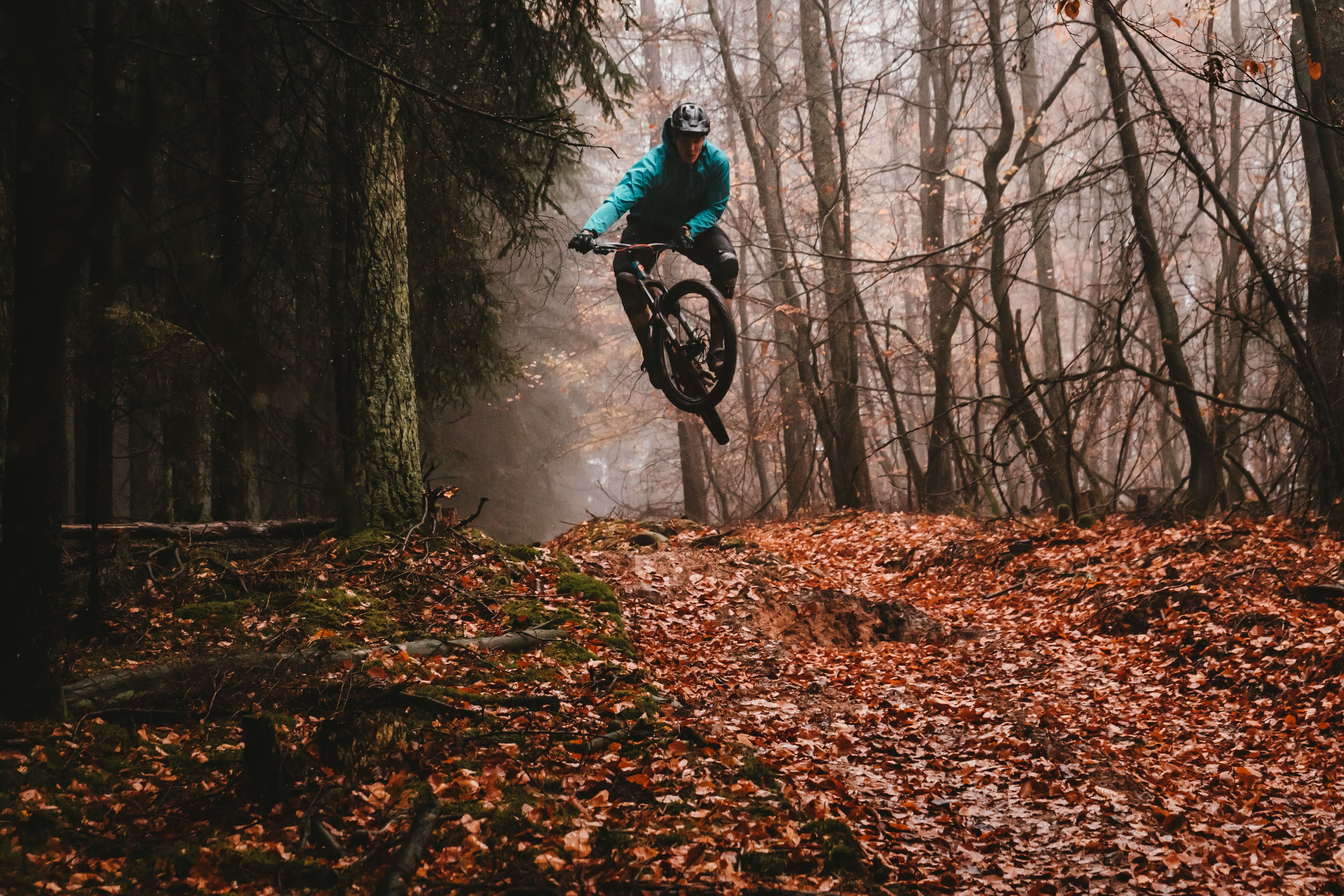 In a misty forest, a man on a mountain bike in turquoise clothing and a bicycle helmet jumps over a hill covered with autumn leaves.