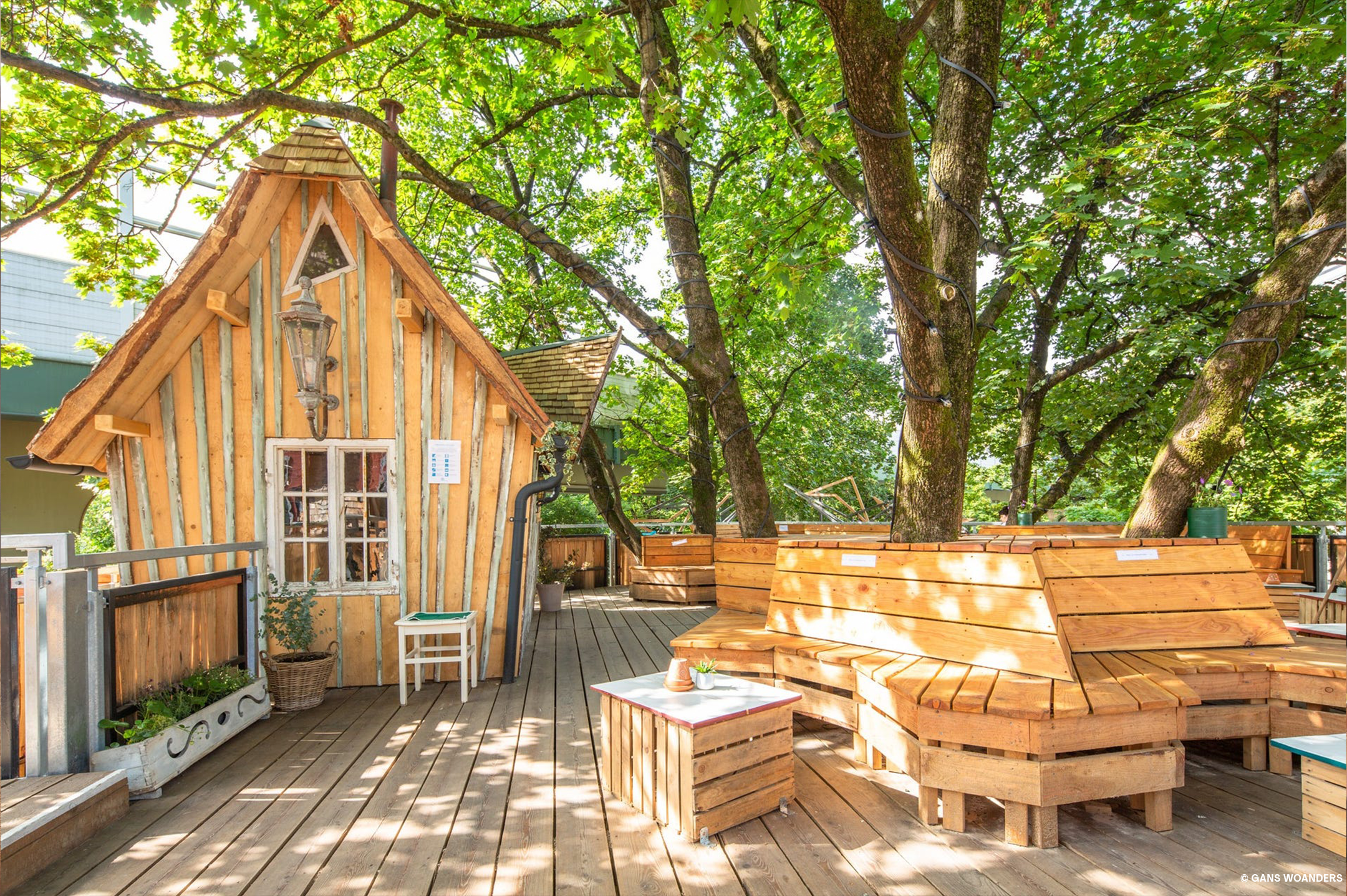 Ein kleines Holzhaus mit direkt anschließender Holzterrasse und Sitzgelegenheiten aus Holz, die Stämme eines Baumes dahinter