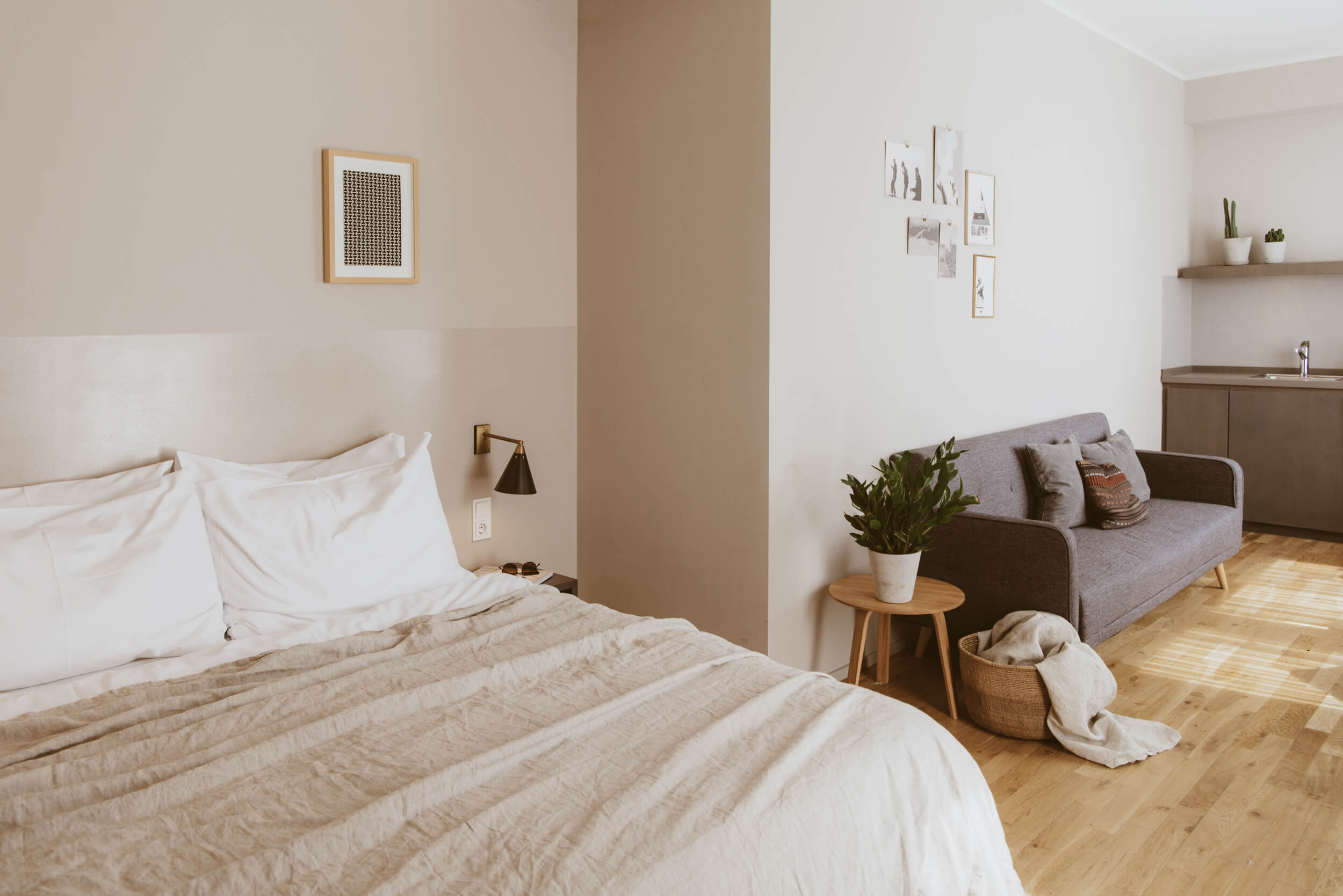 Bold Room im Bold Giesing mit Doppelbett, Sofa und Kitchenette Rechts im Anschnitt ein hell bezogenes Doppelbett, ein graues Sofa mit farbig abgestimmten Kissen an der Wand, rechts dahinter im Anschnitt eine graue Küchenzeile.