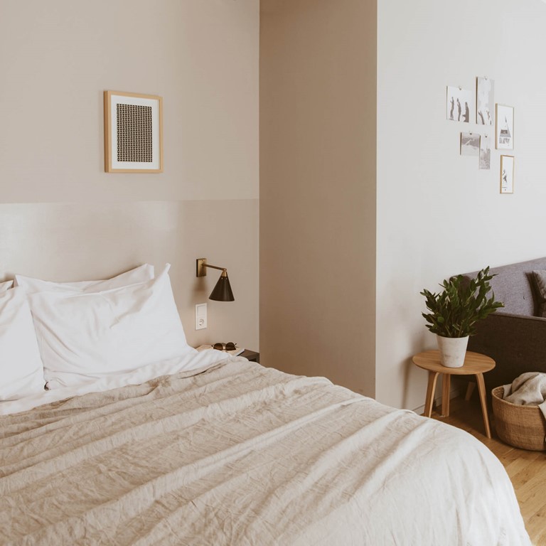 Bold Room im Bold Giesing mit Doppelbett, Sofa und Kitchenette Rechts im Anschnitt ein hell bezogenes Doppelbett, ein graues Sofa mit farbig abgestimmten Kissen an der Wand, rechts dahinter im Anschnitt eine graue Küchenzeile.