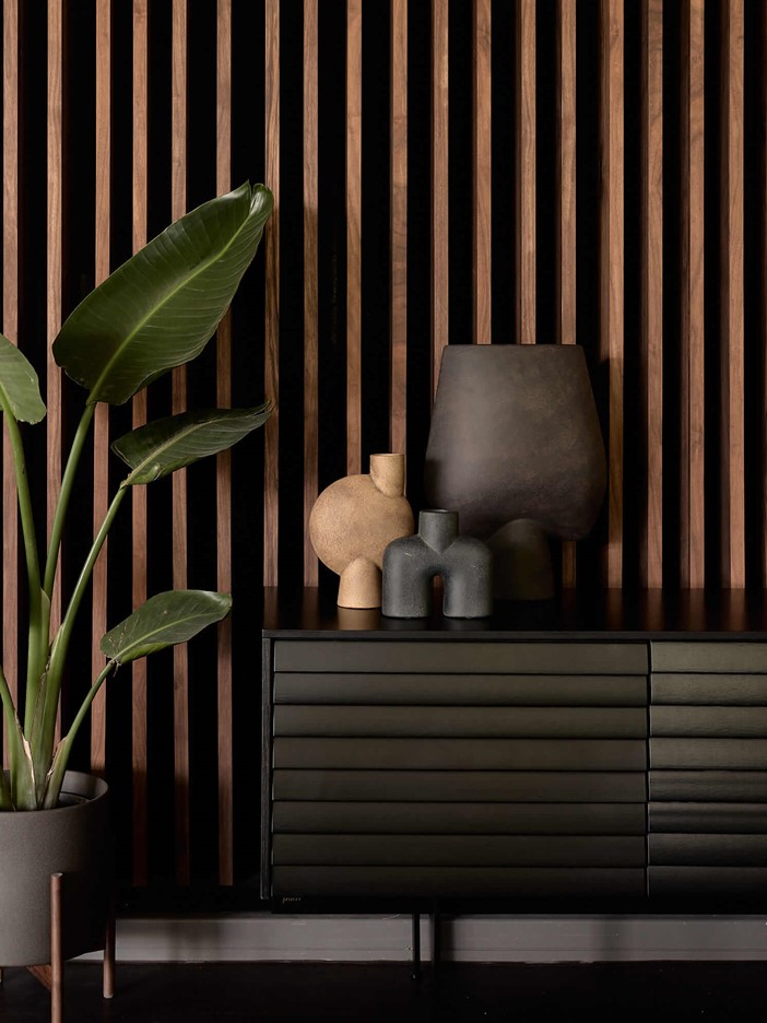 Modern vases on a dark sideboard in front of a screen of wooden elements, a potted plant to the left.